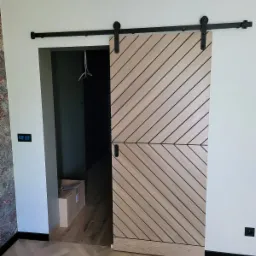 Drewniane drzwi przesuwne w stylu stodoły, z ukośnymi panelami i czarnym systemem przesuwnym, na tle jasnych ścian i parkietu ułożonego w jodełkę.