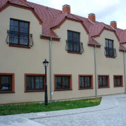 Elewacja budynku wielorodzinnego z rzędami brązowych okien i balkonami z ozdobnymi kratami, widok z poziomu chodnika wyłożonego kostką.