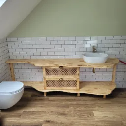 Umywalka nablatowa na blacie z surowego drewna z ażurowymi szufladami w łazience na poddaszu, widoczna toaleta, białe kafelki na ścianie.