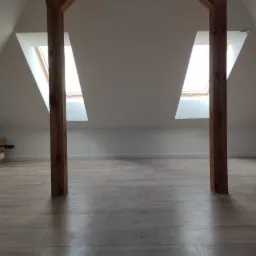 Poddasze po remoncie z dwiema drewnianymi belkami konstrukcyjnymi, dwoma oknami dachowymi i panelami podłogowymi w odcieniach beżu i szarości. W rogu widoczne kartonowe pudełko.