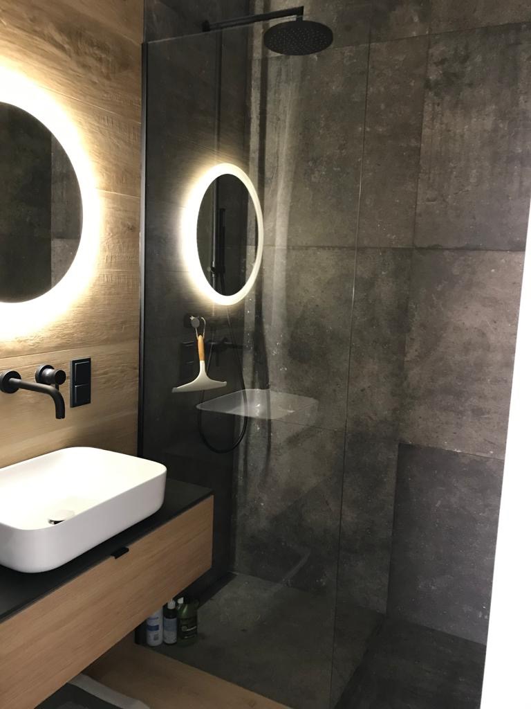 Nowoczesna łazienka z kabiną prysznicową typu walk-in, umywalką nablatową i okrągłym lustrem z podświetleniem LED, wykończona płytkami w odcieniach szarości i drewna.