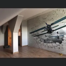 Pokój na poddaszu z drewnianą podłogą, szarą ścianą z fototapetą 3D przedstawiającą samoloty przebijające się przez mur z cegieł, ukośne ściany i drewniane drzwi.