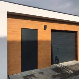 Nowoczesny, parterowy budynek z bramą garażową i ciemnymi drzwiami wejściowymi, wykończony w odcieniach bieli i brązu z dekoracyjną strukturą na elewacji, oświetlony bocznym światłem słonecznym.