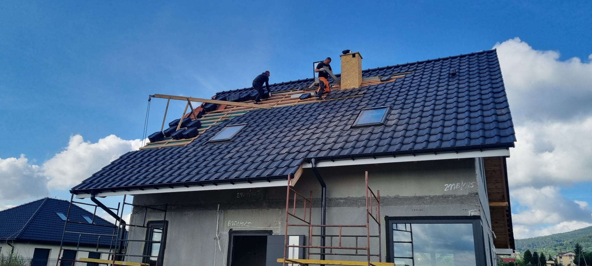 Montaż czarnej dachówki na nowym domu z oknami dachowymi, widoczni dekarze pracujący na dachu, na dole rusztowania.