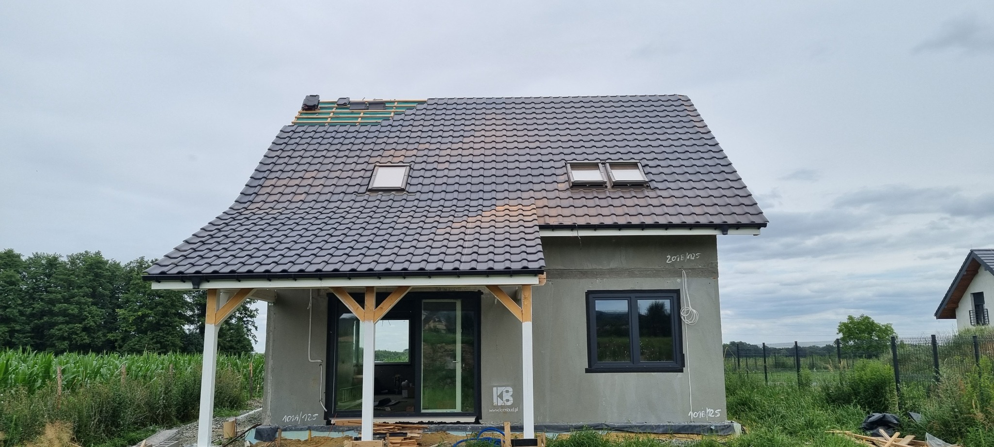 Dom w trakcie krycia dachu z widocznymi łatami, częściowo ułożoną dachówką w kolorze grafitowym i oknami dachowymi, elewacja w szarym tynku, z przodu zadaszenie z drewnianymi słupami.