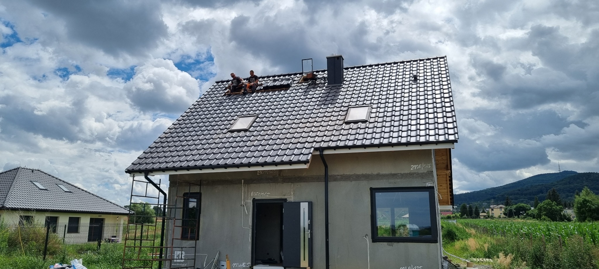 Dwóch dekarzy pracuje na dachu domu jednorodzinnego pokrytym ciemną dachówką, widoczne okna dachowe i komin, w tle pochmurne niebo i pobliskie budynki.
