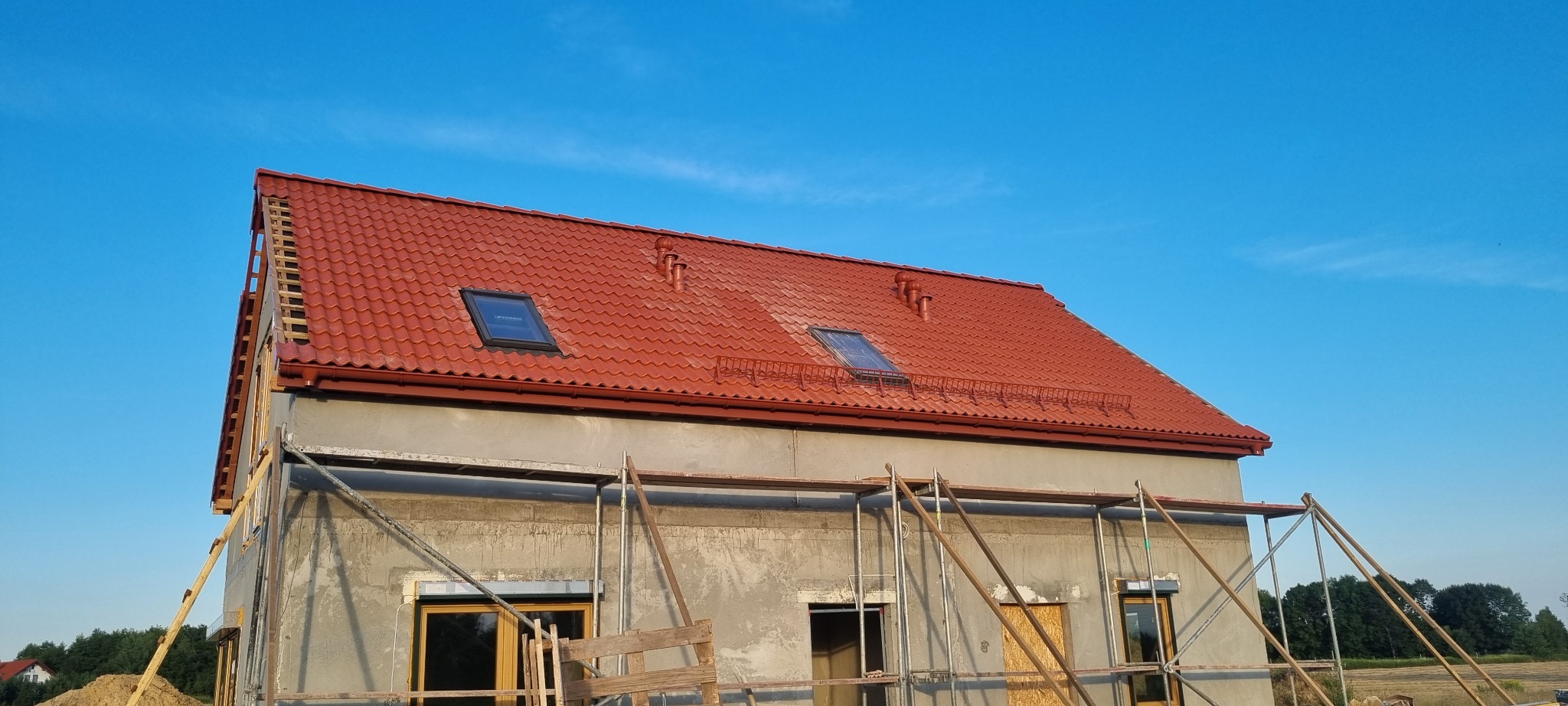 Dom w trakcie budowy z nowym, czerwonym dachem z dachówki ceramicznej i oknami dachowymi, widoczne rusztowania i elementy więźby dachowej na krawędzi dachu.