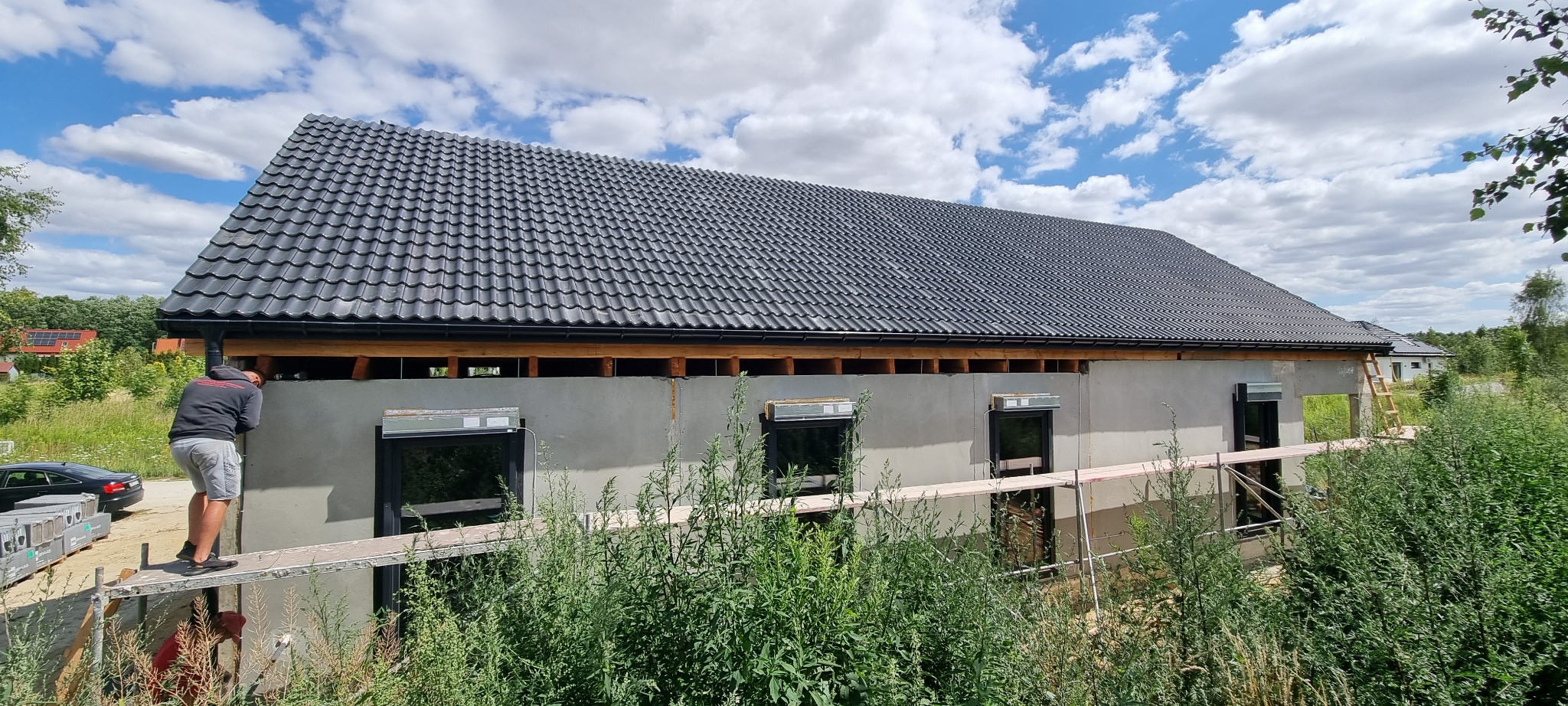 Czarny dach z dachówki ceramicznej na domu w trakcie budowy; widoczna elewacja w stanie surowym, rusztowanie i pracownik wykonujący prace wykończeniowe na wysokości