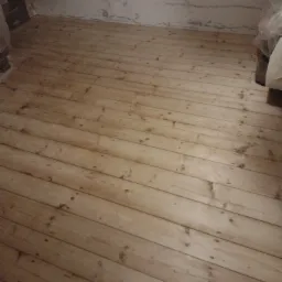 Jasna, drewniana podłoga z widocznymi śladami po cyklinowaniu, ściany w tle przygotowane do malowania, widoczne niedoskonałości i ubytki.