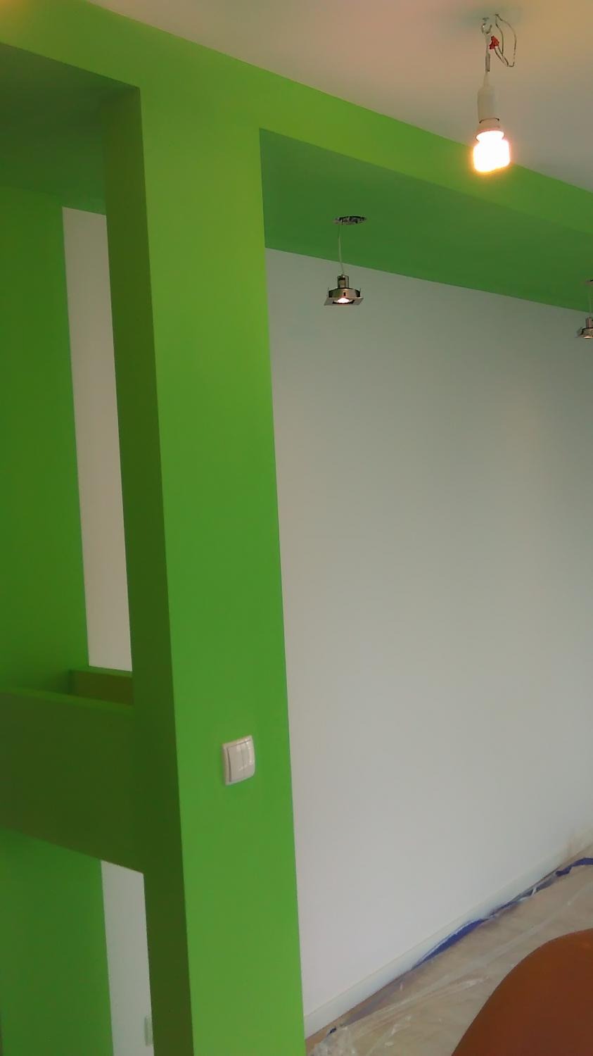 Jasnozielona ściana z dekoracyjnymi elementami architektonicznymi i włącznikiem światła, kontrastująca z białą ścianą i trzema lampami sufitowymi. Podłoga zabezpieczona folią malarską.