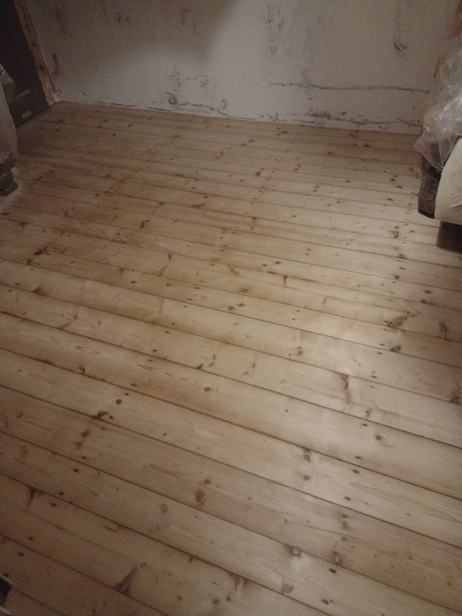 Jasna, drewniana podłoga z widocznymi śladami po cyklinowaniu, ściany w tle przygotowane do malowania, widoczne niedoskonałości i ubytki.