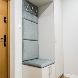 Meble Kułakowski Grzegorz Kułakowski - Wnęka z siedziskiem i wieszakami w przedpokoju, obok drzwi wejściowych, biała zabudowa meblowa z szafkami, logo firmy na górze.