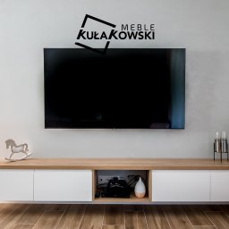 Meble Kułakowski Grzegorz Kułakowski - Nowoczesna szafka RTV na wymiar z białymi frontami i drewnianym blatem, dekorowana drewnianym konikiem na biegunach i szklanymi wazonami, pod wiszącym telewizorem z logo firmy Kułakowski Meble...