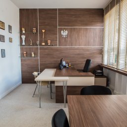 Meble Kułakowski Grzegorz Kułakowski - Biuro z drewnianą zabudową, biurkami i regałami, ozdobione godłem Polski i trofeami, logo firmy meblarskiej w rogu kadru.