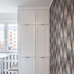 Meble Kułakowski Grzegorz Kułakowski - Biała szafa wnękowa z czterema frontami z minimalistycznymi uchwytami, dopasowana do szarej podłogi i geometrycznej tapety, widok z boku, z oknem w tle.