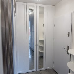 Meble Kułakowski Grzegorz Kułakowski - Biała szafa wnękowa z lustrzanymi drzwiami w przedpokoju, widoczny fragment wieszaka i półek w środku, nad szafą kryształowy żyrandol, na górze logo firmy.