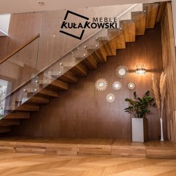 Meble Kułakowski Grzegorz Kułakowski - Drewniane schody z szklaną balustradą, oświetlone lampą ścienną i dekorowane lustrami w stylu boho, z logo firmy 'MEBLE KUŁAKOWSKI' w górnym rogu.