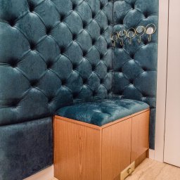 Meble Kułakowski Grzegorz Kułakowski - Turkusowy, pikowany panel ścienny z siedziskiem obitym tą samą tkaniną, uzupełniony szafką z jasnego drewna i złotymi wieszakami, całość w eleganckim stylu.