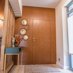 Meble Kułakowski Grzegorz Kułakowski - Nowoczesny przedpokój z drewnianą zabudową ściany i ukrytymi drzwiami, dekoracyjnymi lustrami w złotych ramach, zielonym stolikiem z kwiatami i dywanem w geometryczny wzór.