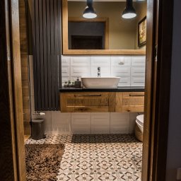 Meble Kułakowski Grzegorz Kułakowski - Nowoczesna łazienka z drewnianymi meblami na wymiar, białą umywalką nablatową i wzorzystą podłogą, widok zza drewnianej framugi drzwiowej, logo producenta mebli widoczne w odbiciu lustra.