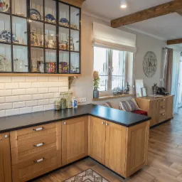 Jasna kuchnia z drewnianymi szafkami, czarnym blatem i przeszkloną witryną wypełnioną ceramiką, wzorzysta podłoga, w tle okno i komoda, logo firmy w lewym górnym rogu.