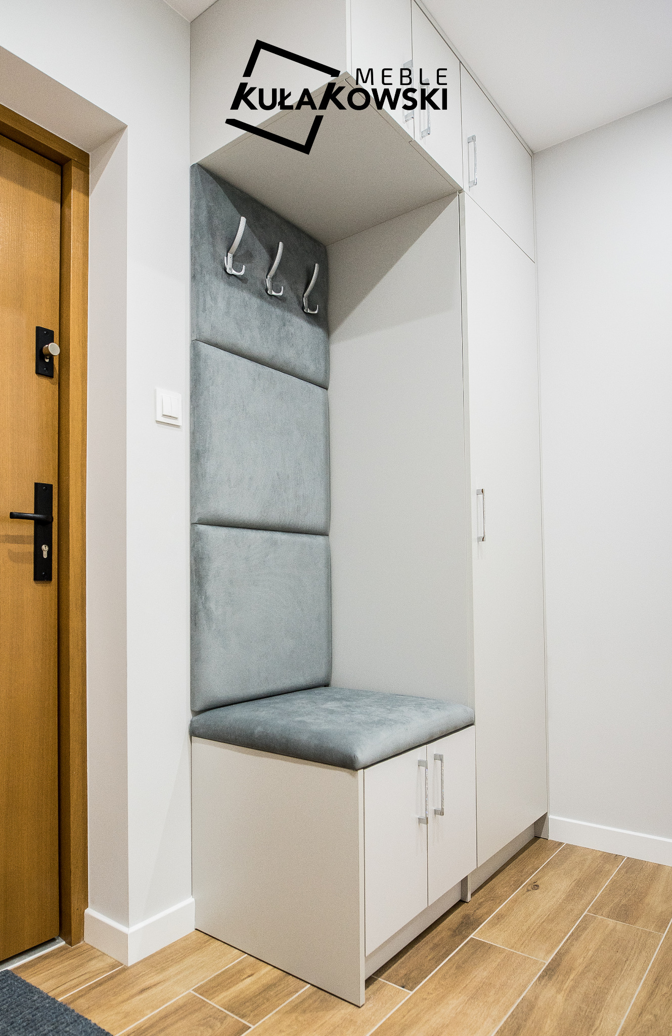 Wnęka z siedziskiem i wieszakami w przedpokoju, obok drzwi wejściowych, biała zabudowa meblowa z szafkami, logo firmy na górze.