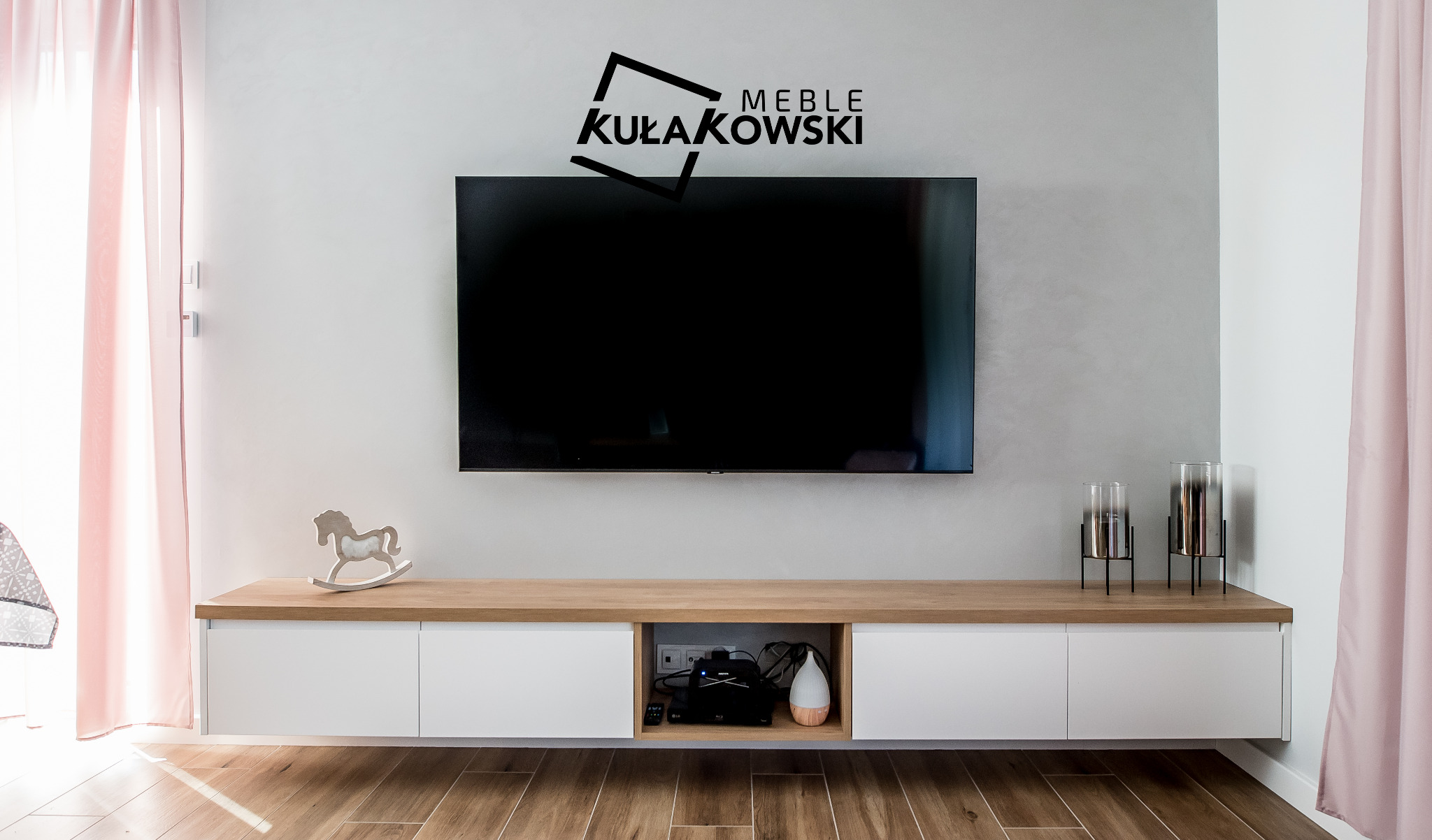 Nowoczesna szafka RTV na wymiar z białymi frontami i drewnianym blatem, dekorowana drewnianym konikiem na biegunach i szklanymi wazonami, pod wiszącym telewizorem z logo firmy Kułakowski Meble...