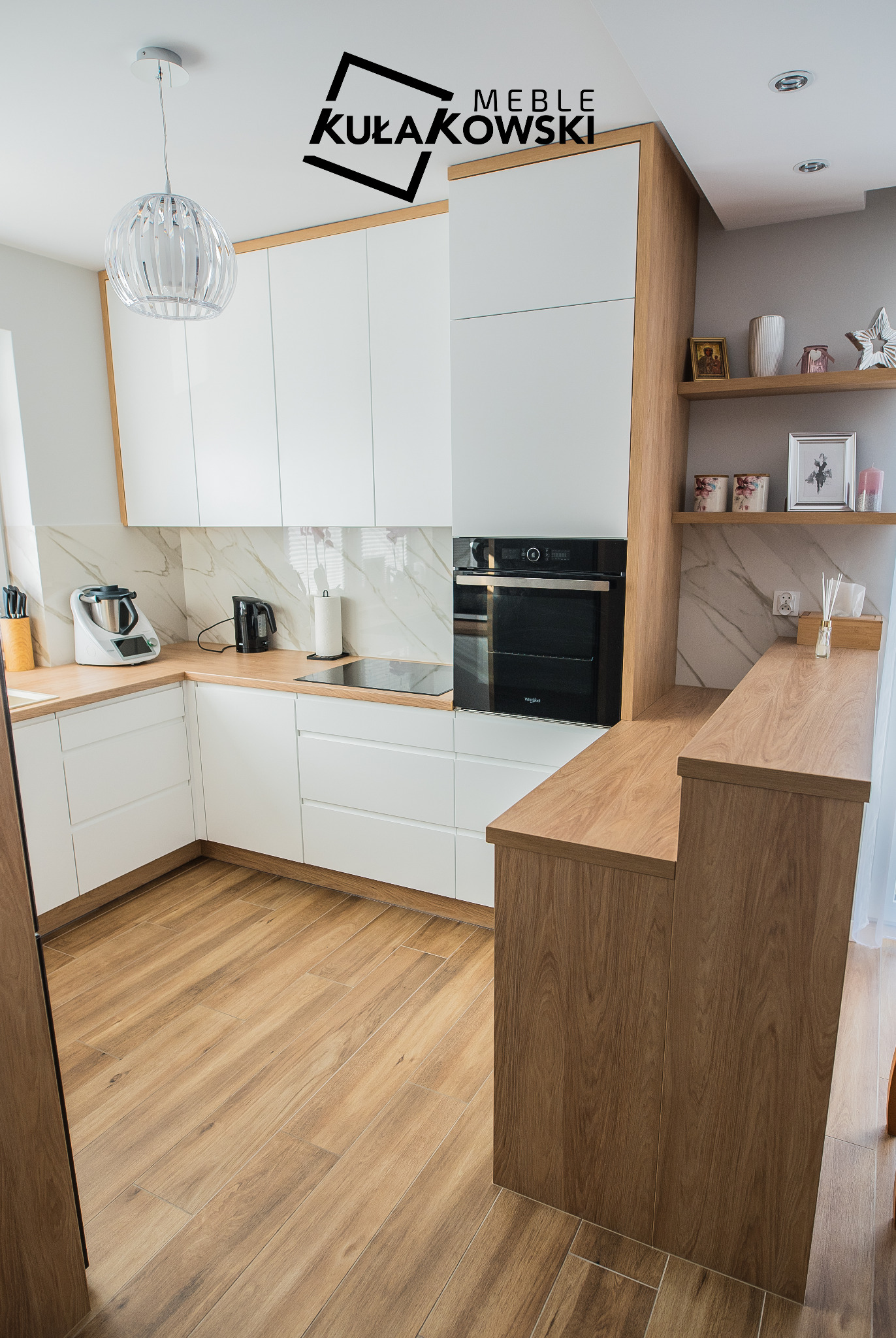 Nowoczesna kuchnia z białymi frontami i drewnianymi blatami, widoczny sprzęt AGD oraz dekoracyjne półki z dodatkami, oświetlona wiszącą lampą i z logo firmy w górnej części kadru.