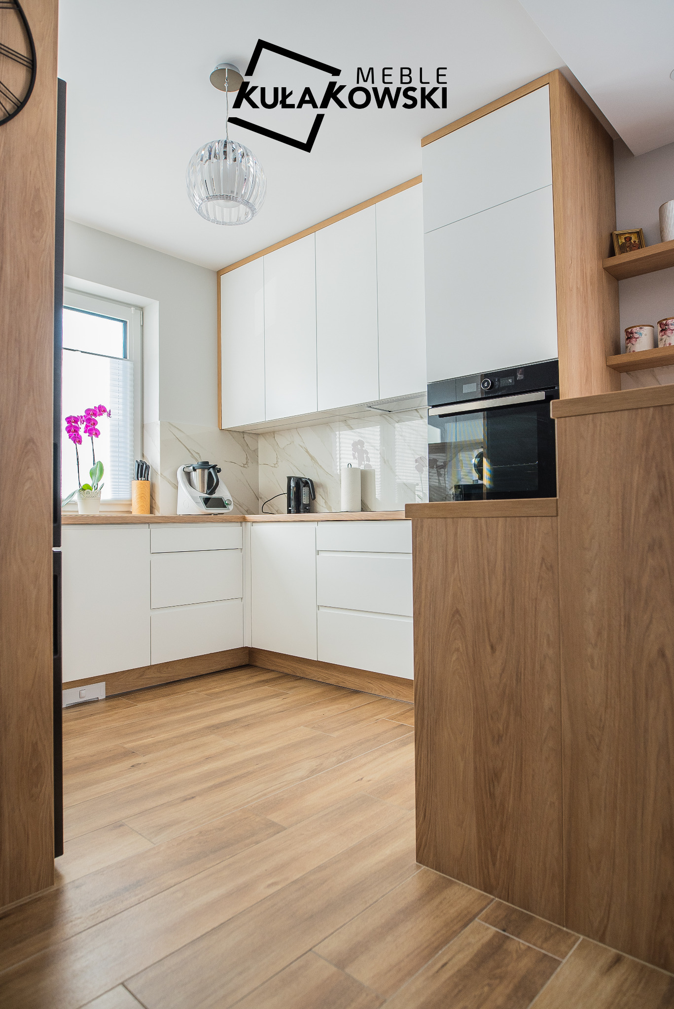 Jasna kuchnia z białymi szafkami i drewnianymi akcentami, widok z perspektywy wejścia, na blacie sprzęt AGD i doniczka z kwiatami, na ścianie logo firmy.