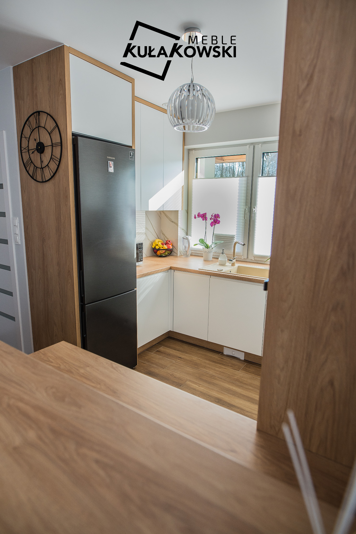 Jasna kuchnia z białymi szafkami, drewnianym blatem i podłogą, czarną lodówką i okrągłym zegarem na ścianie, oświetlona ozdobną lampą wiszącą, z widocznym logo firmy na suficie.