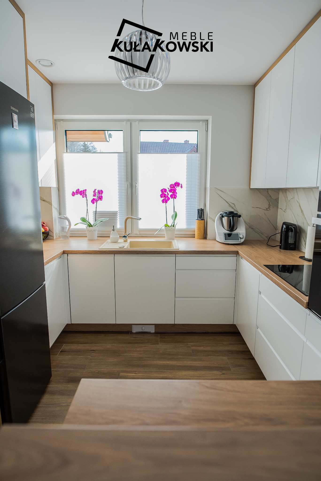 Jasna kuchnia w stylu skandynawskim z białymi frontami i drewnianymi blatami, z widocznym oknem z roletami plisowanymi i dwoma storczykami w doniczkach, czarna lodówka Samsung po lewej stronie...