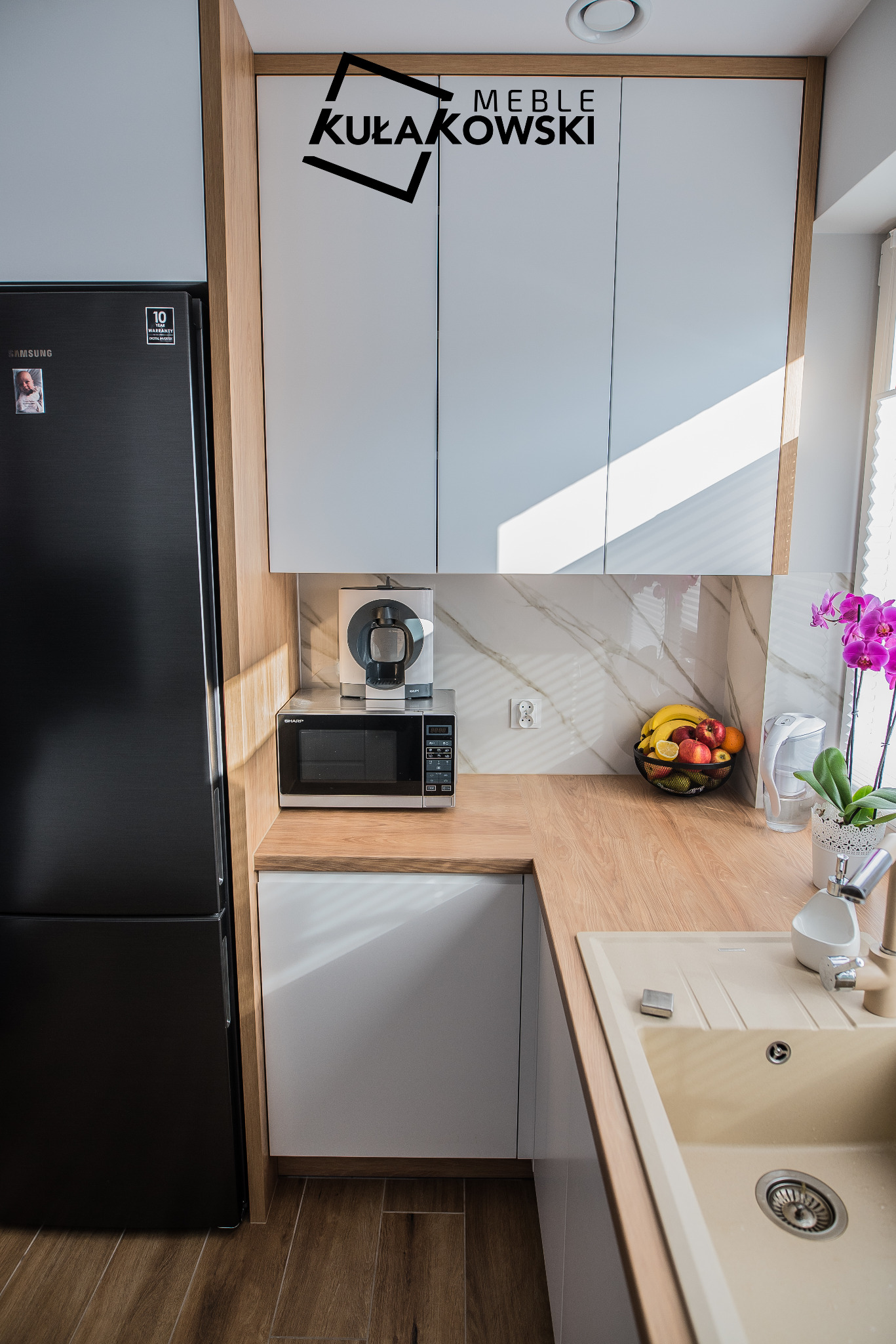 Nowoczesna kuchnia z czarną lodówką Samsung, białymi szafkami z drewnianymi akcentami, blatem w kolorze drewna, zlewem w odcieniach beżu, mikrofalówką Sharp i ekspresem do kawy Krups, oświetlona...