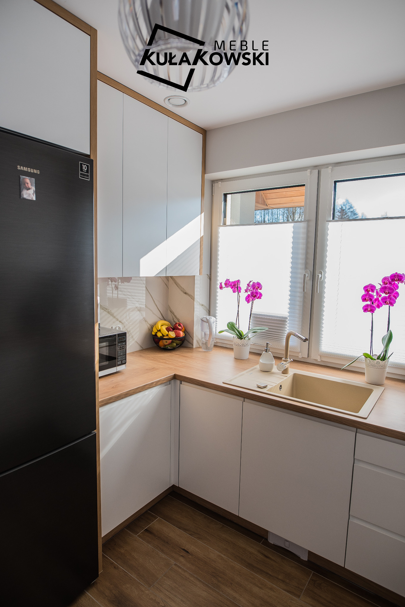 Nowoczesna kuchnia z białymi szafkami, drewnianym blatem i podłogą, czarną lodówką Samsung, zlewozmywakiem w kolorze piaskowym, dwoma fioletowymi orchideami w doniczkach na parapecie okna...
