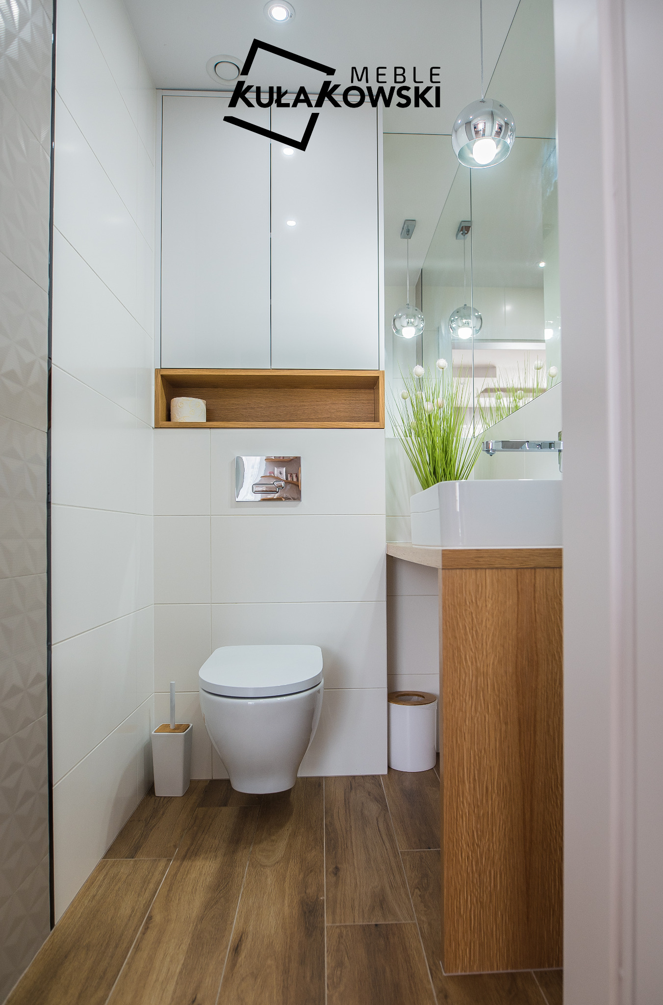 Nowoczesna łazienka z drewnianą podłogą, białymi płytkami, podwieszaną toaletą, drewnianą półką, umywalką na drewnianym blacie i lustrem z wiszącymi lampami, oświetlona punktowymi światłami,...