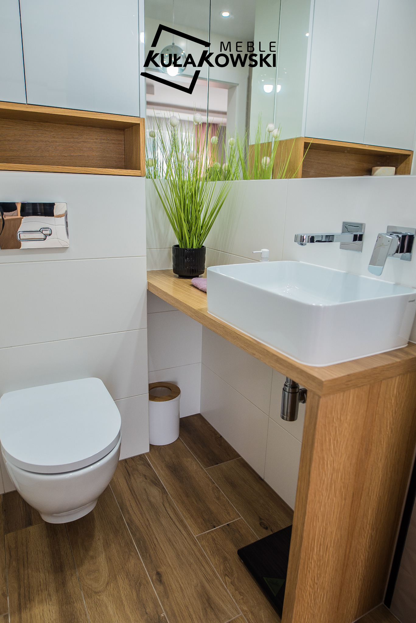 Nowoczesna łazienka z drewnianym blatem pod umywalką, białą ceramiką i drewnopodobną podłogą, w lustrze odbija się logo firmy meblarskiej.