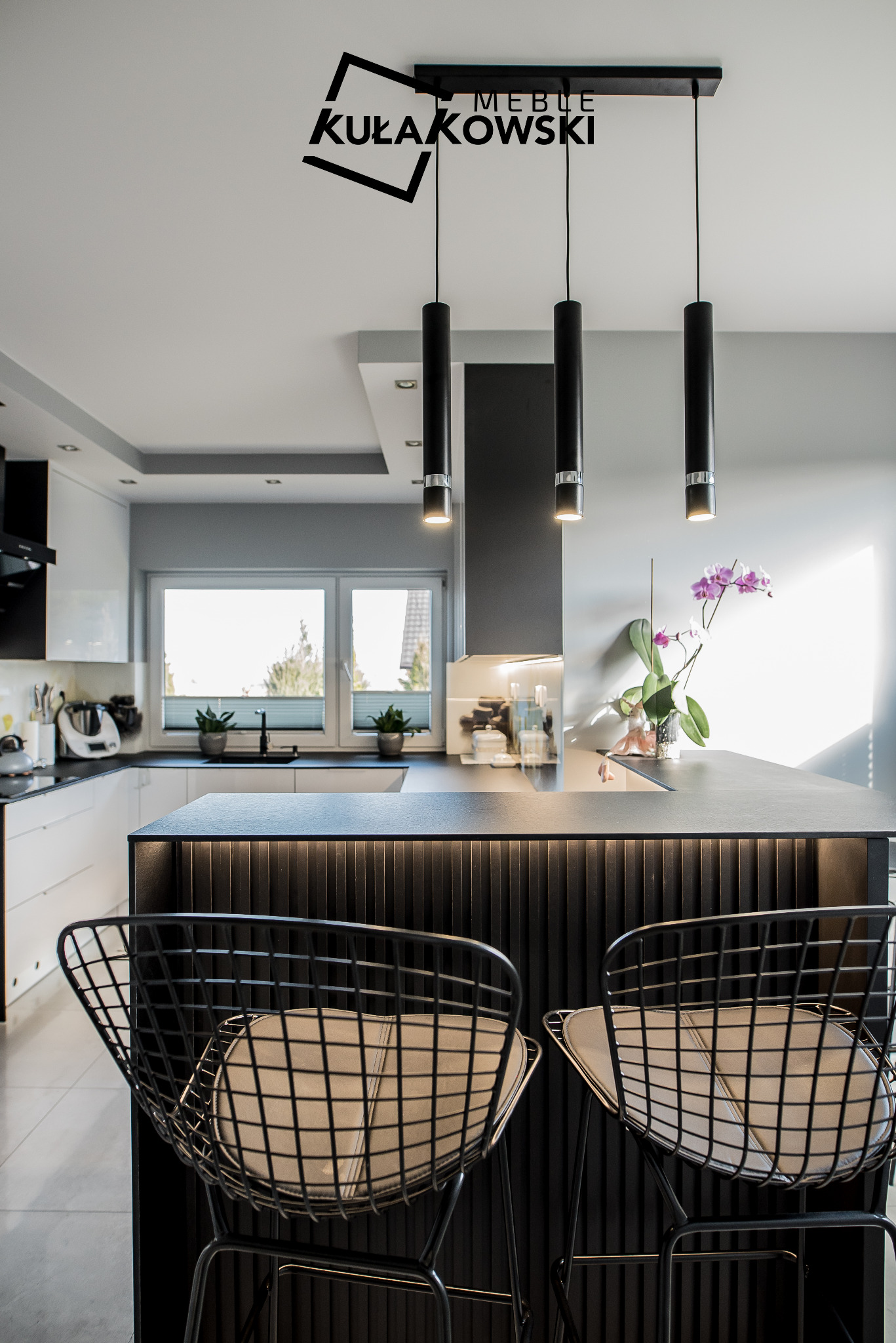 Nowoczesna kuchnia z czarnym blatem wyspowym z podświetlanymi panelami, czarne metalowe hokery z jasnymi poduszkami, wiszące lampy cylindryczne i białe szafki w tle, ozdobiona storczykiem.