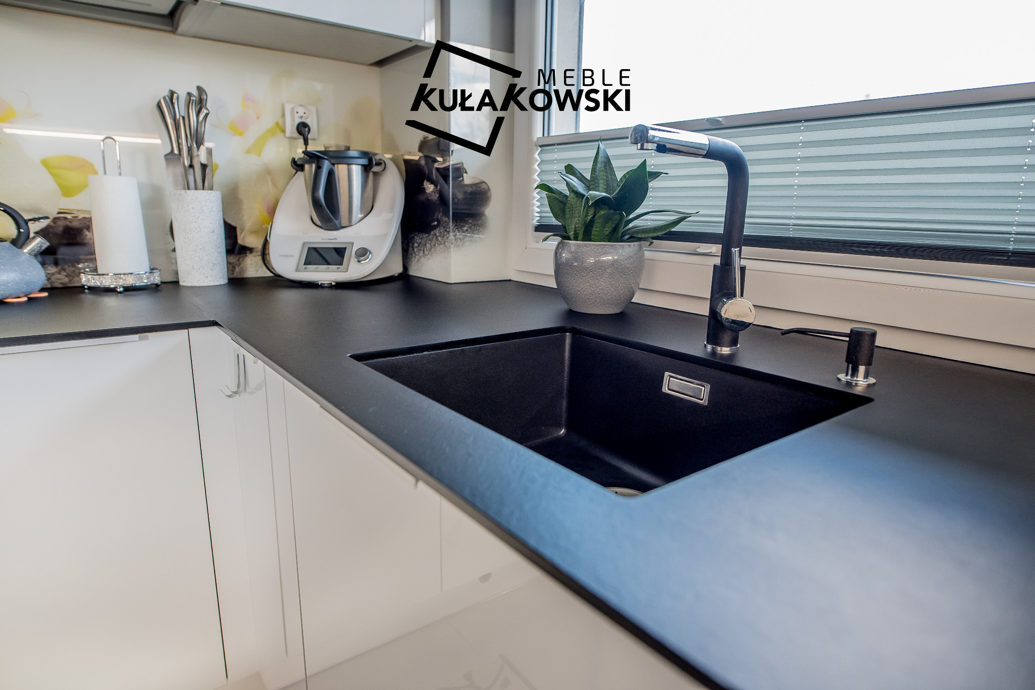 Czarny, matowy zlew wpuszczony w czarny blat, bateria kuchenna w kolorze chromu, białe szafki kuchenne i dekoracyjny panel ścienny z motywem kwiatowym w tle.