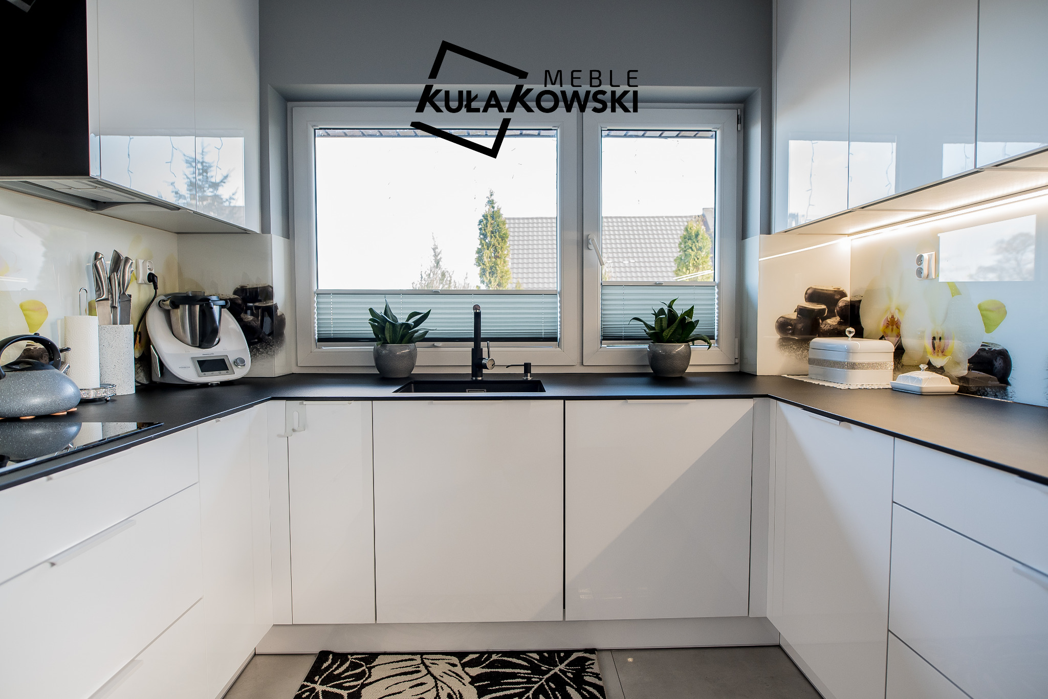 Nowoczesna, biała kuchnia z czarnym blatem i minimalistycznym designem, z oknem między szafkami górnymi a blatem, ozdobiona dwoma doniczkami z roślinami i dywanikiem w liście.
