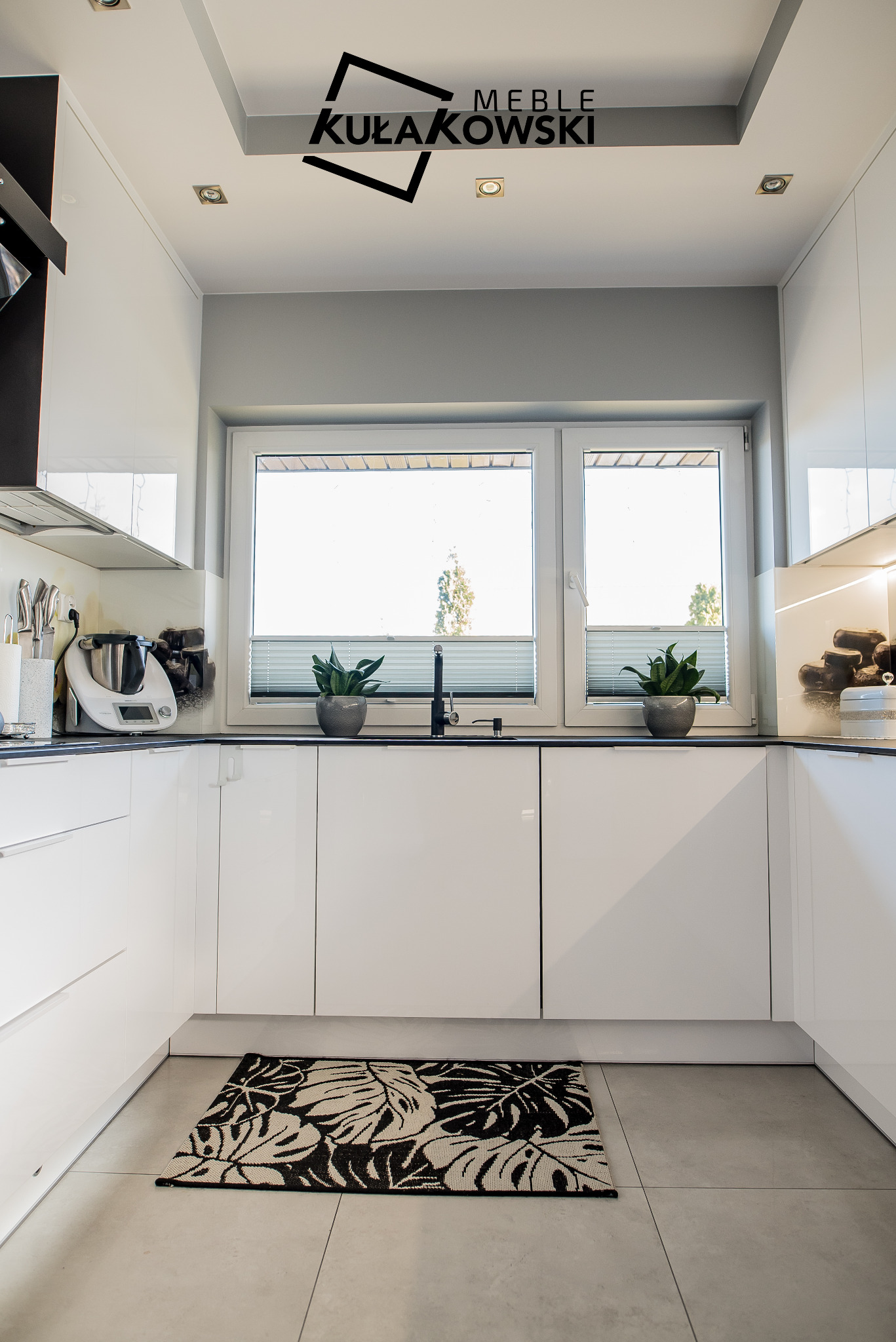 Biała kuchnia na wymiar z czarnym blatem, roślinami w doniczkach przy oknie i dywanem w liście na podłodze; logo firmy widoczne na suficie.