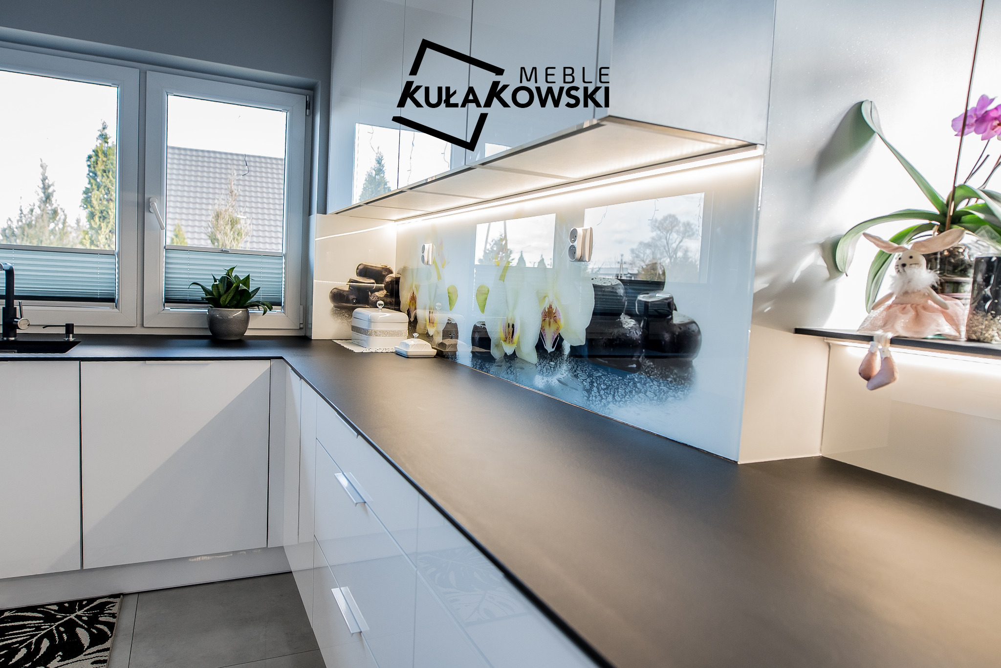Nowoczesna kuchnia z białymi, lakierowanymi frontami, czarnym blatem i szklanym panelem z motywem storczyków, oświetlonym od góry. Widoczne logo firmy meblarskiej nad panelem.