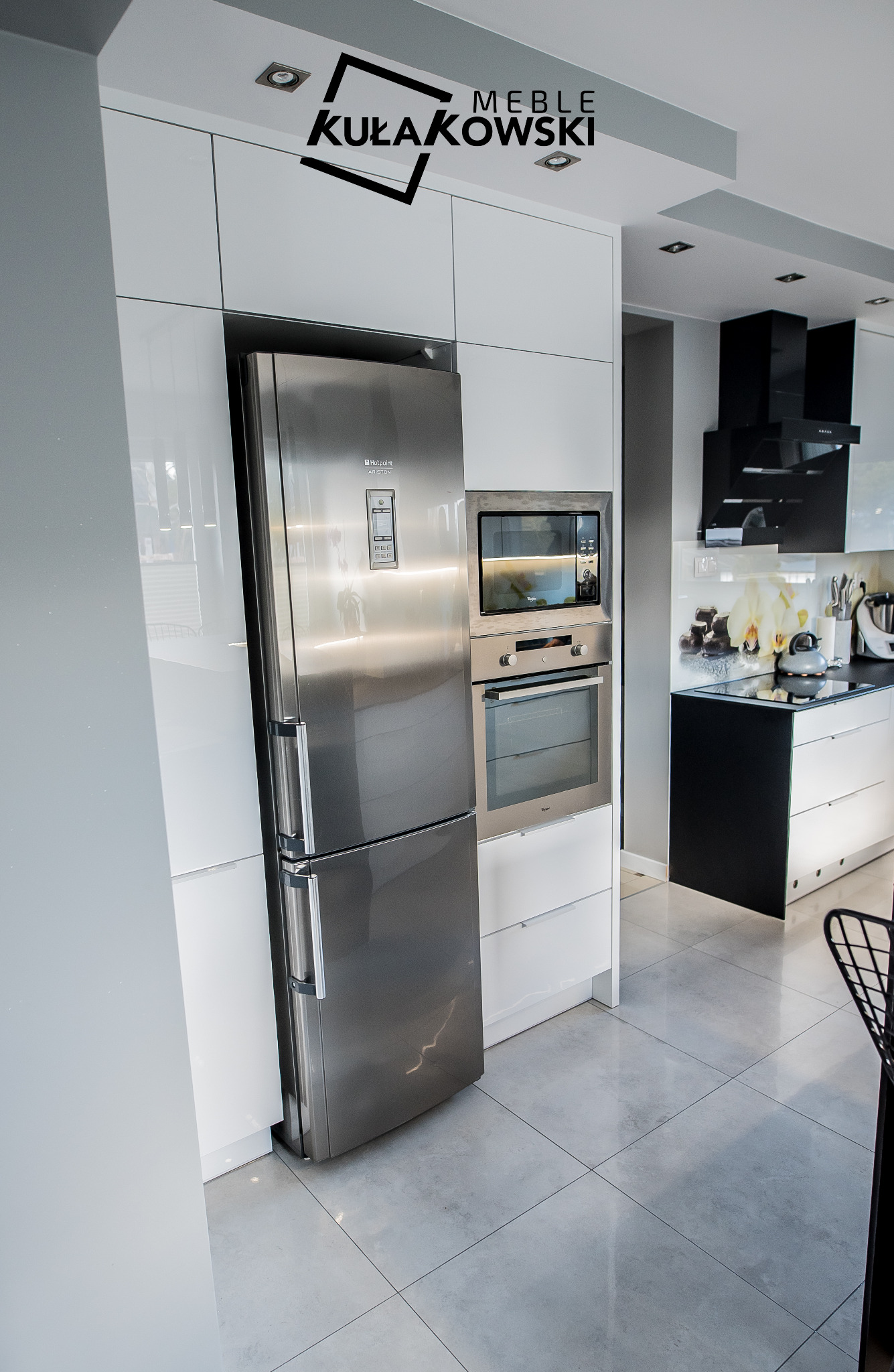 Nowoczesna, biała kuchnia z połyskiem, z zabudowaną lodówką ze stali nierdzewnej i piekarnikiem, widoczny fragment logo firmy meblowej na górze.