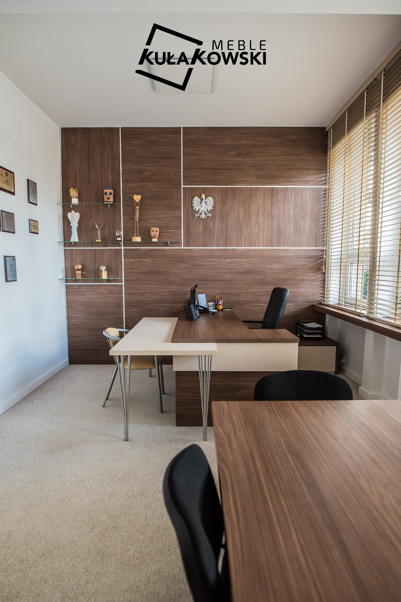 Biuro z drewnianą zabudową, biurkami i regałami, ozdobione godłem Polski i trofeami, logo firmy meblarskiej w rogu kadru.