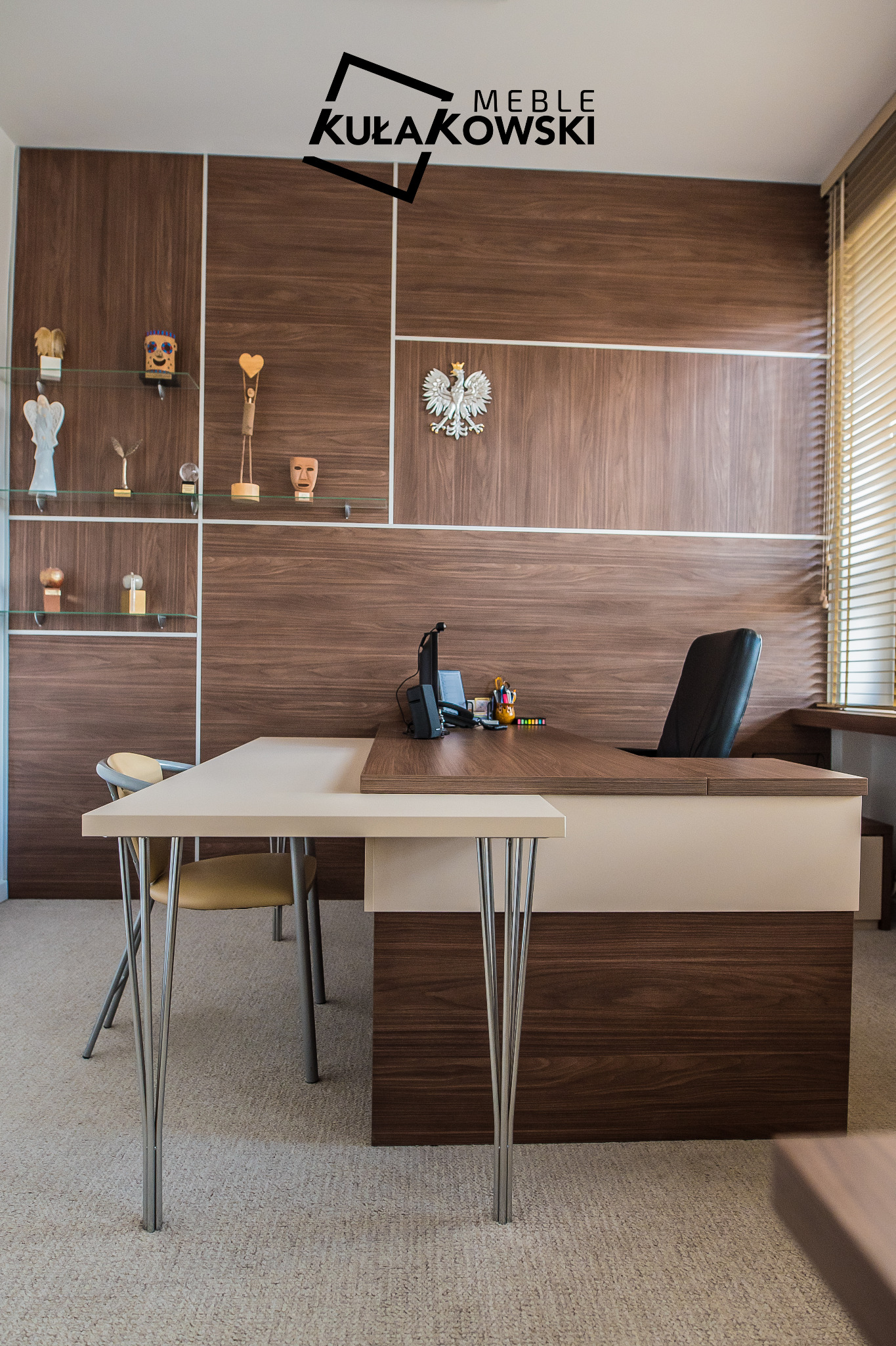Gabinet z biurkiem z forniru orzechowego, krzesłami na metalowych nogach i wbudowanymi półkami z dekoracjami, na ścianie godło państwowe, logo firmy meblarskiej w rogu.