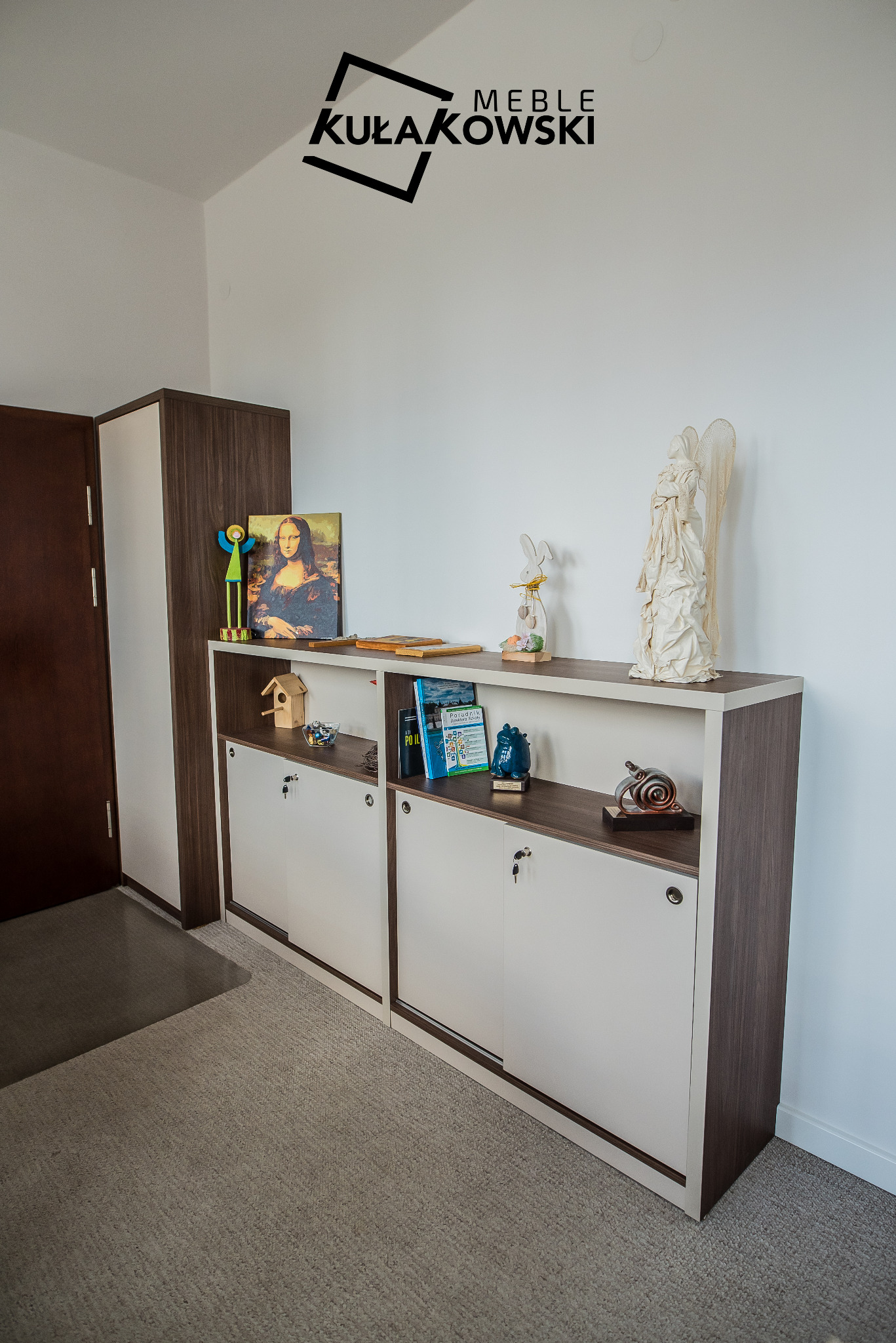 Dwudrzwiowa szafka z przesuwnymi drzwiami w kolorze białym i ciemnego drewna, z otwartymi półkami, na których stoją dekoracje, w tle logo firmy meblowej.