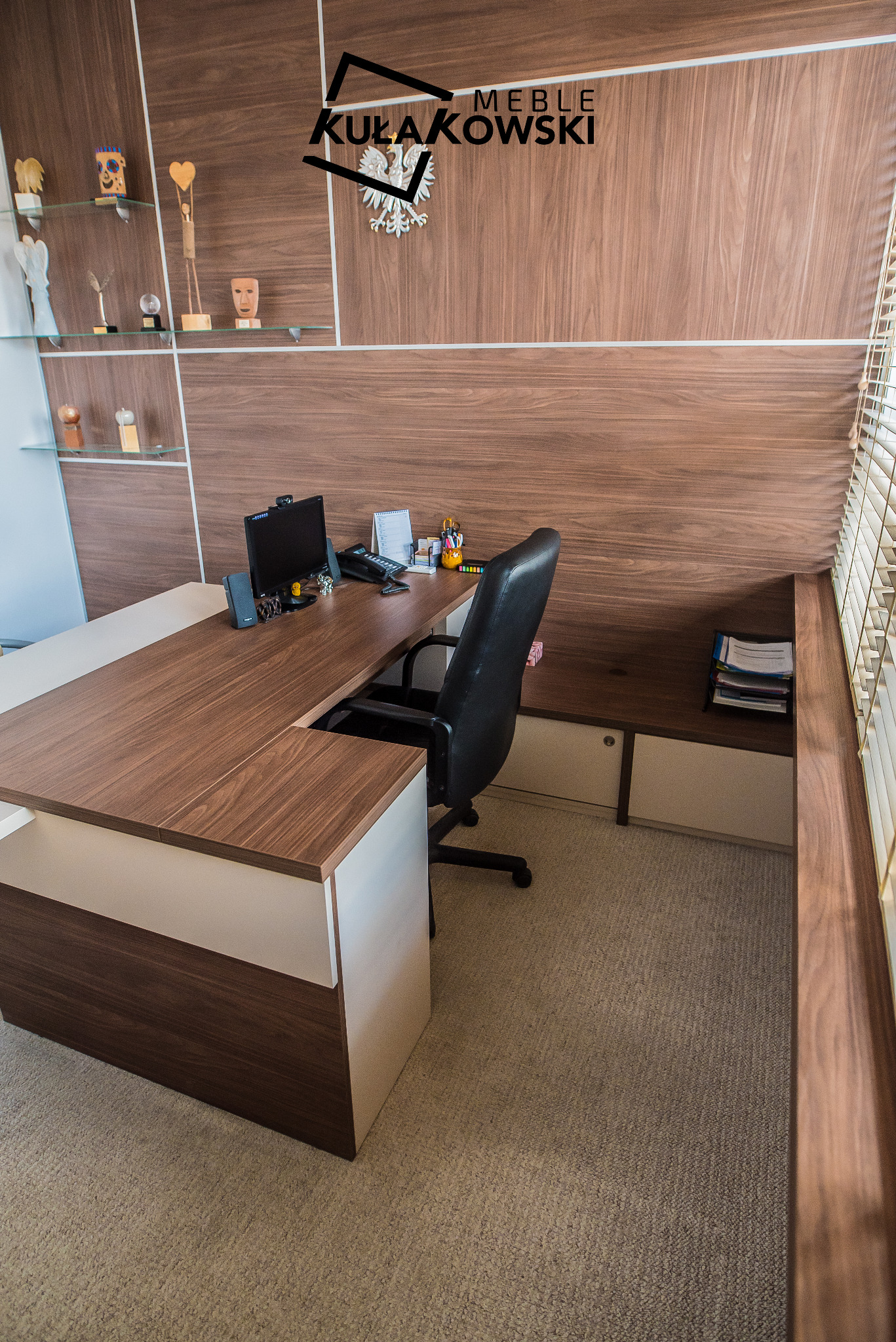 Biuro z drewnianym biurkiem na wymiar, zabudową ścienną z półkami i siedziskiem, czarnym krzesłem obrotowym i widocznym logo firmy na ścianie.