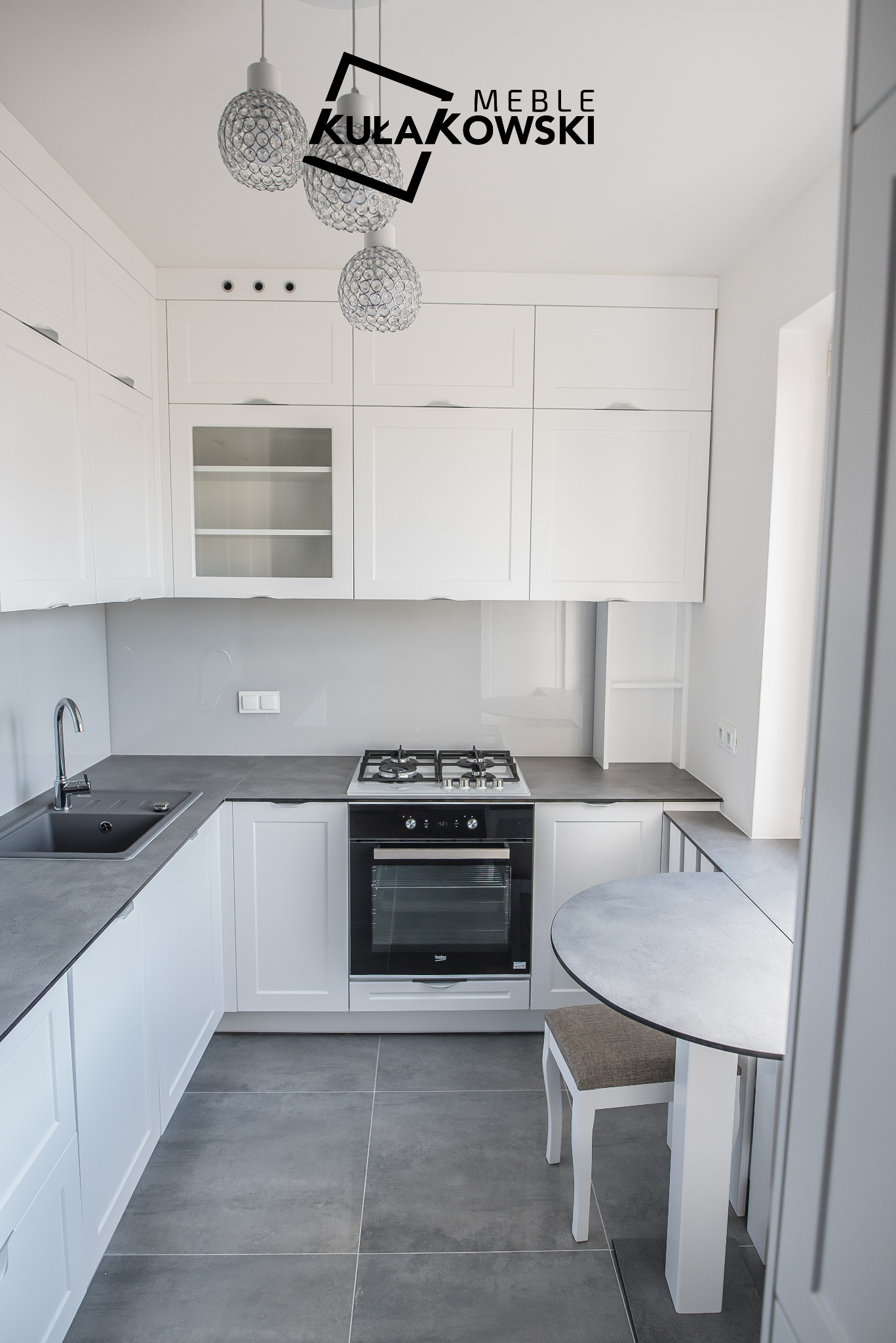 Jasna kuchnia na wymiar z szarymi blatami i podłogą, oświetlona ozdobnymi lampami, z widocznym logo producenta mebli.
