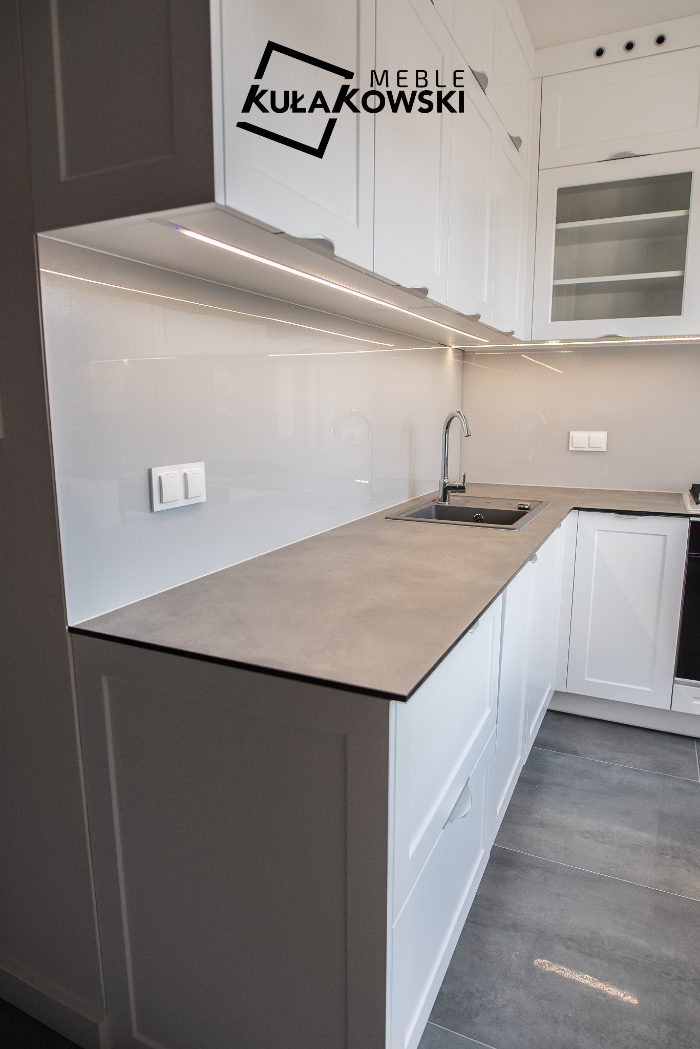 Nowoczesna, jasna kuchnia z białymi szafkami, szarym blatem i szklanym panelem ściennym podświetlonym listwą LED. Widoczne logo firmy 'Meble Kułakowski' na górnych szafkach.
