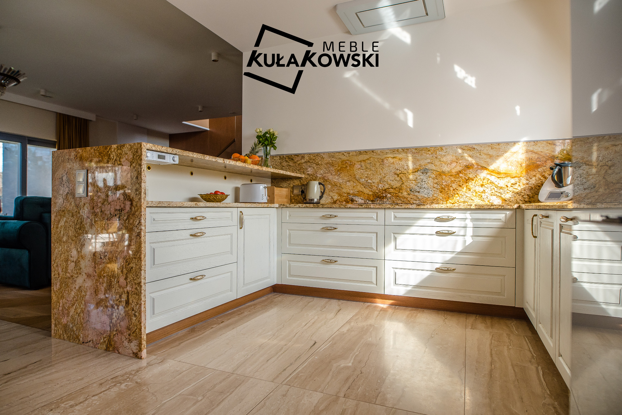 Jasna kuchnia z białymi szafkami i blatem z kamienia w odcieniach złota i brązu, widoczny sprzęt AGD i dekoracje, logo firmy na ścianie, podłoga z płytek imitujących marmur.