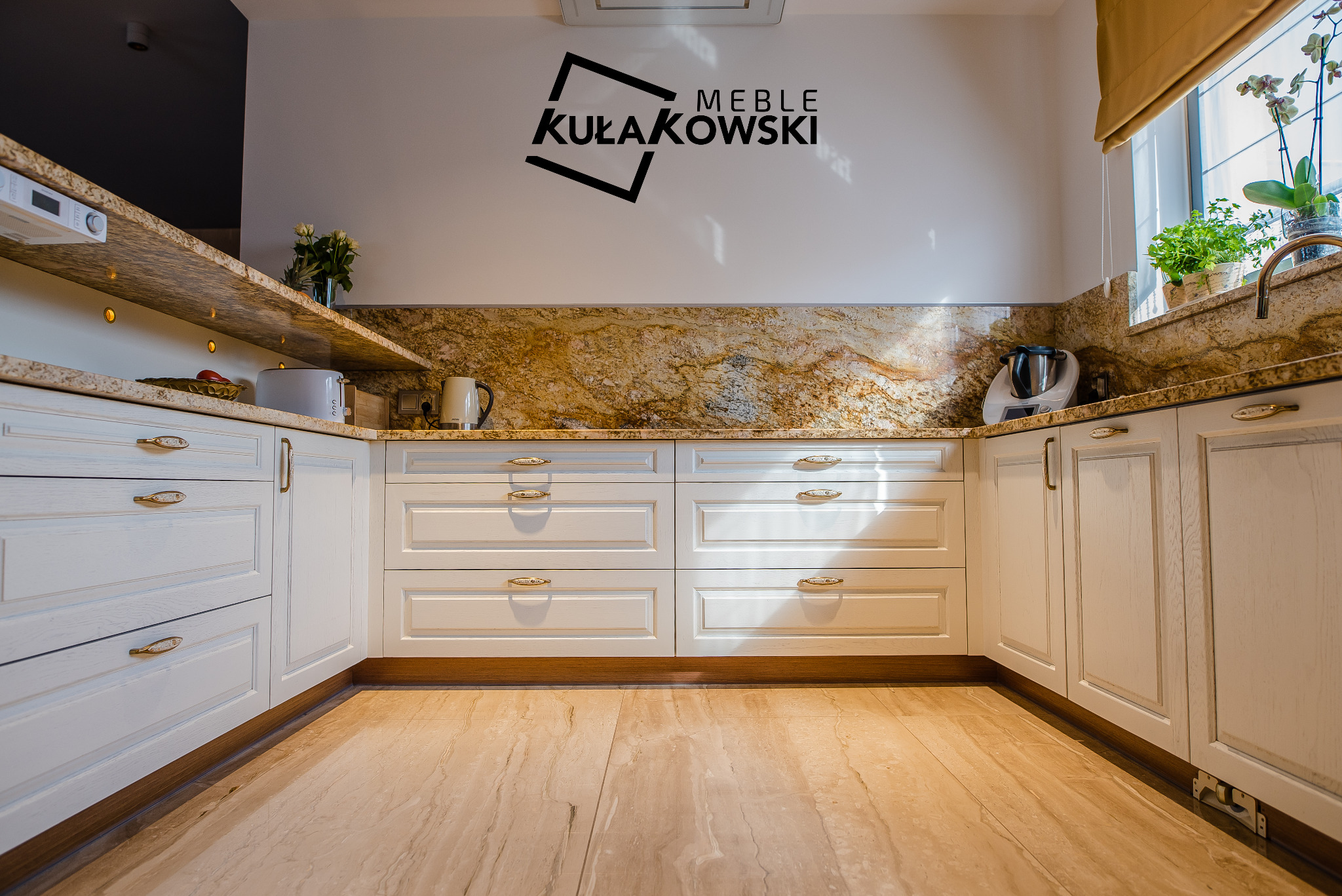 Jasna kuchnia z białymi szafkami z frezowanymi frontami i złotymi uchwytami, blatem z kamienia w odcieniach brązu i beżu oraz podłogą z płytek imitujących kamień. Na ścianie logo firmy 'Meble...