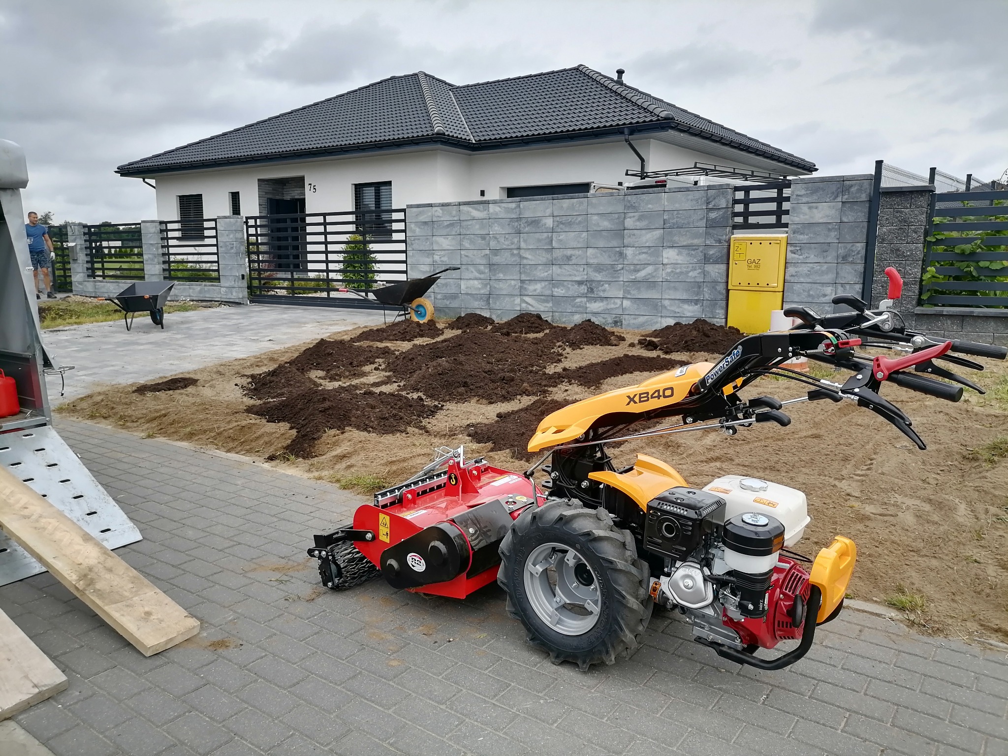 Żółto-czerwony glebogryzarkowy traktor XB40 na kostce brukowej przed domem z szarą kostką elewacyjną i czarnym ogrodzeniem, przygotowujący teren pod trawnik.